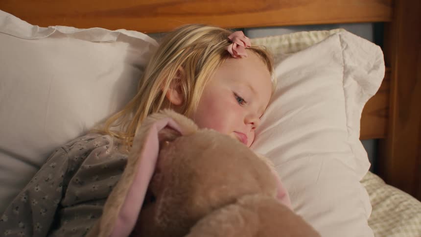 A sleepy little blonde girl in her pajamas holds her stuffed bunny close as she lies in bed, ready for a nap or bedtime