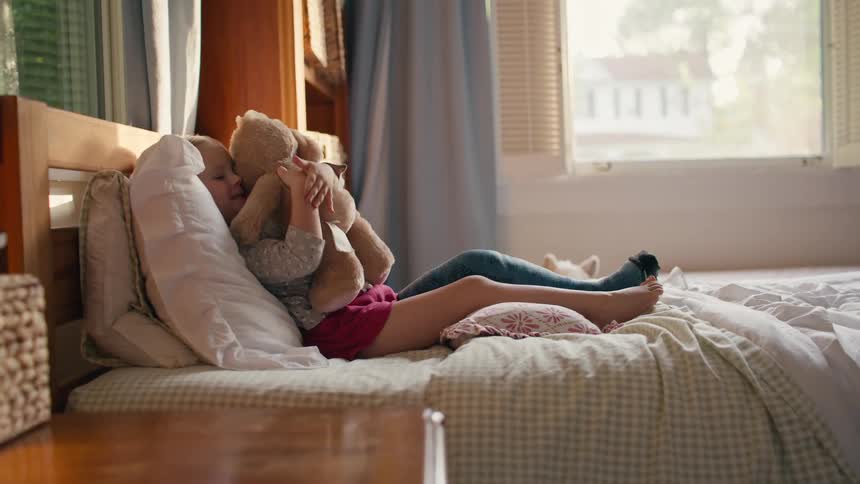 Young girl relaxes on a cozy bed cuddling a stuffed bunny in a sunlit bedroom