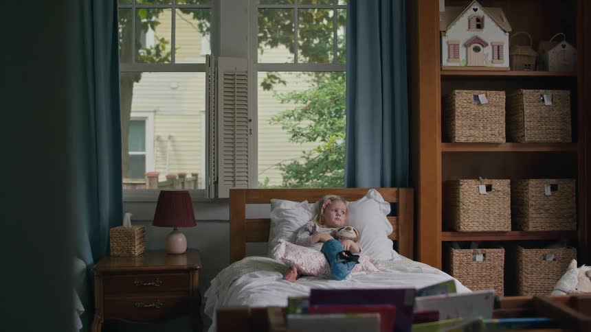 A young girl with a stuffed animal lays in her bed watching something off-screen in her bedroom