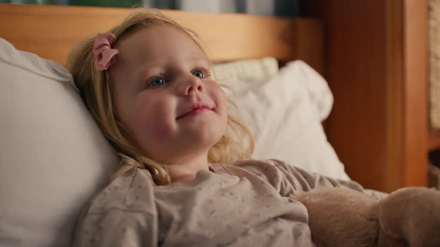 Close up of a happy toddler with blue eyes and a pink bow in her hair smiling contentedly while resting in bed with her teddy bear