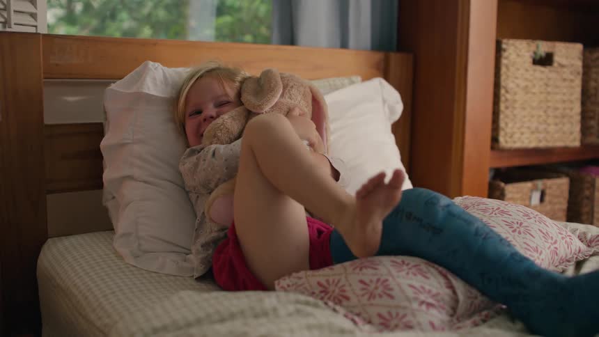 Happy little girl with a cast on her leg rests in bed while smiling and cuddling her stuffed bunny rabbit