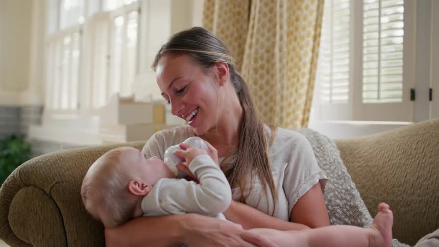 A loving mother smiles as she bottle-feeds her baby while sitting on the couch in their sunlit living room