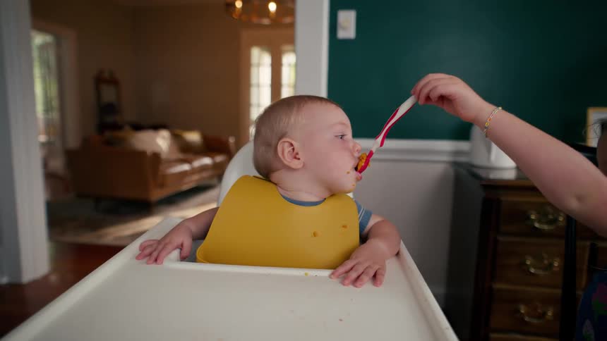 A cute toddler girl carefully feeding her baby brother at the dinner table in their home