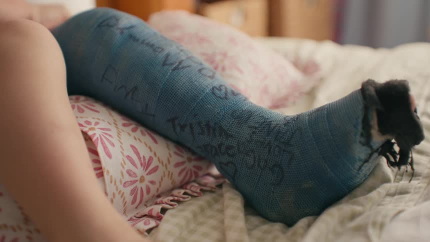 A close-up shot of a person's leg in a blue cast covered with signatures and messages, resting on pillows in bed