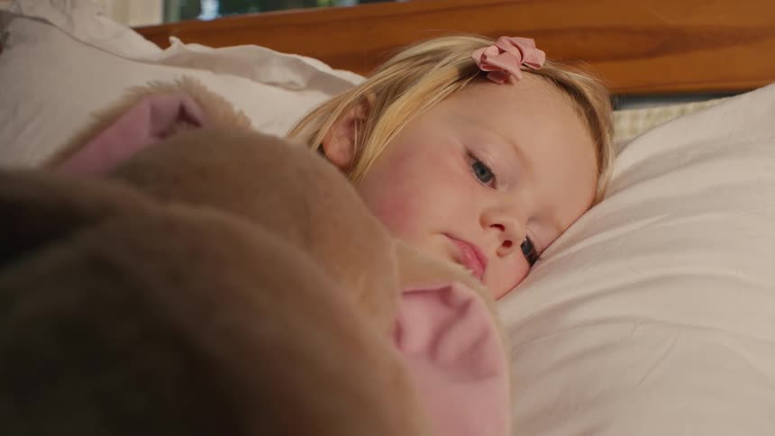 Young blonde girl lies awake in bed cuddling a stuffed animal with a quiet, thoughtful expression