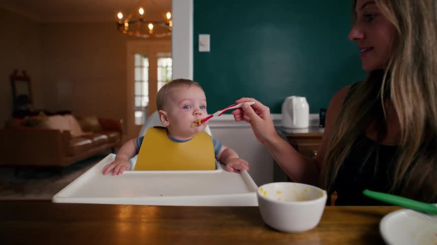 Mother feeding her baby boy with a spoon as he sits in a high chair at the dining table