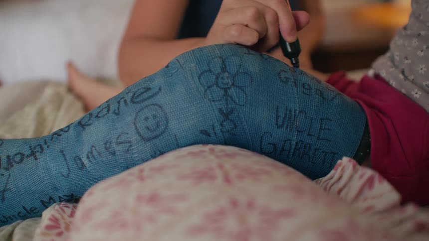 A friend signs their name with a black marker on a young person's blue leg cast already covered with signatures