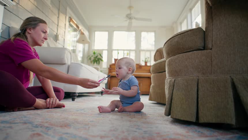 A mother sits on the floor and spoon-feeds her cute baby boy who then takes the spoon to chew on it