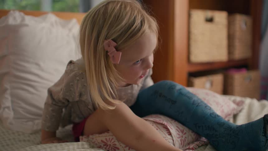 A cute little girl with a cast on her leg sits up in bed and smiles happily while watching something off-screen