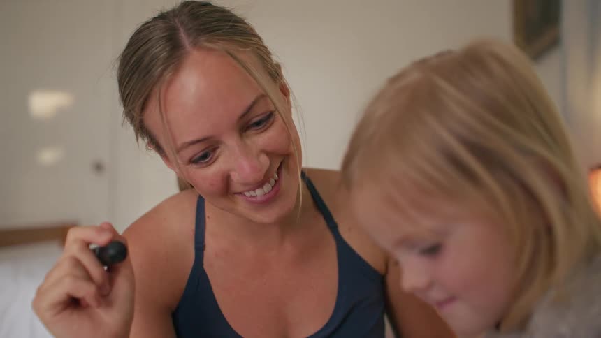A close-up shot captures a loving mother smiling as she draws with her young daughter in a cozy room