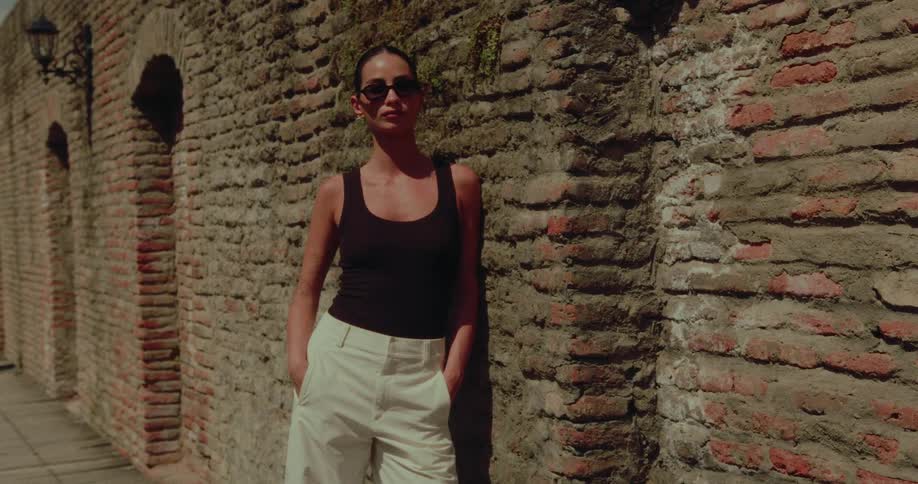A stylish woman in sunglasses poses confidently against a rustic brick wall on a sunny day