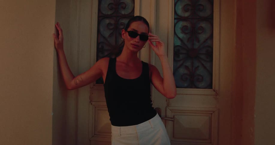 A stylish woman in sunglasses strikes a confident pose in a warmly lit, elegant doorway at night