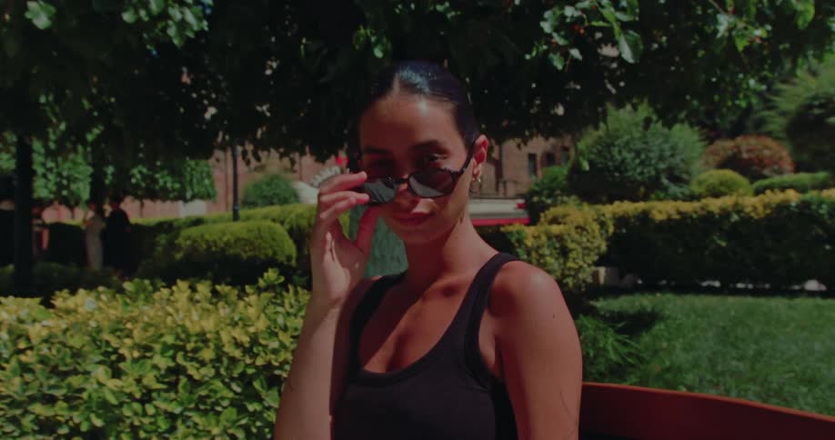 A beautiful brunette lowers her sunglasses to give a mysterious smile while sitting on a bench in a lush green park