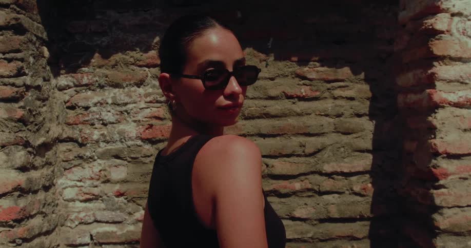 A stylish woman in sunglasses turns to look over her shoulder while standing against an old brick wall in the sun