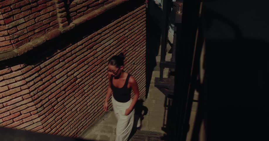 From a high angle, a stylish woman in sunglasses walks up a narrow stone staircase in a sunlit historic alley