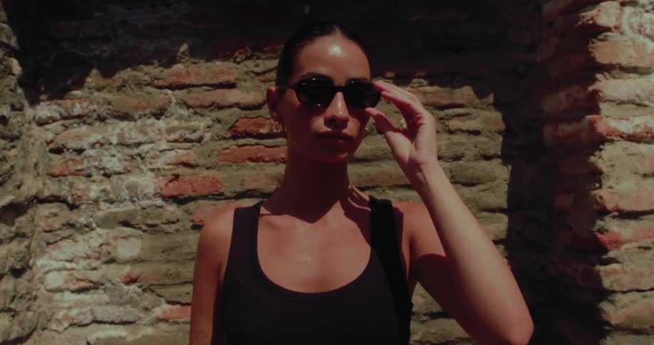 A stylish woman in sunglasses adjusts them in slow motion while standing against a rustic brick wall on a sunny day
