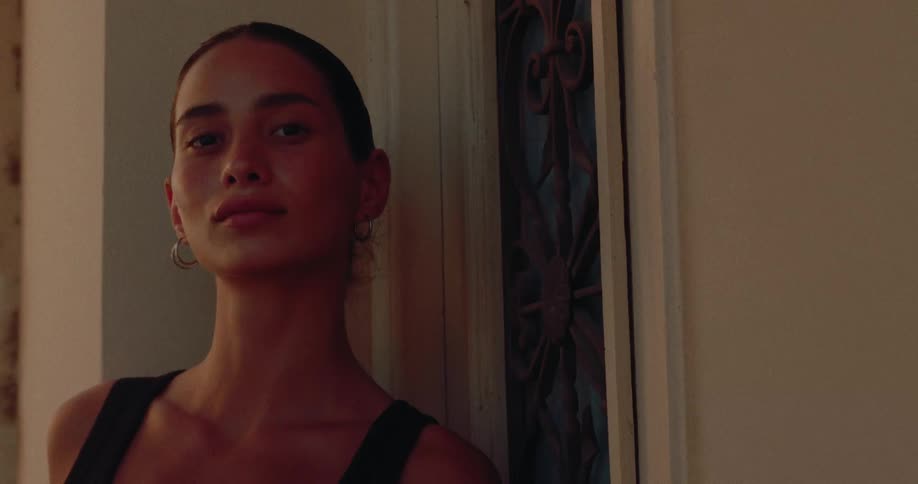 A close-up portrait of a pensive young woman with her hair pulled back looking directly at the camera in a warmly lit, moody setting