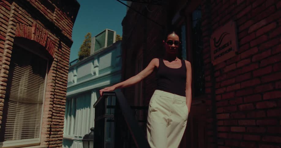 A stylish woman in sunglasses poses on a stoop in a sunny, narrow brick alleyway