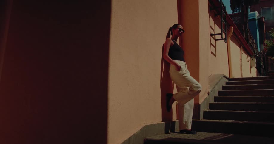 A stylish young woman in sunglasses leans casually against a sunlit wall next to a city staircase