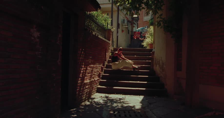 A woman sits on sunlit stairs in a narrow, shadowy alleyway on a sunny day