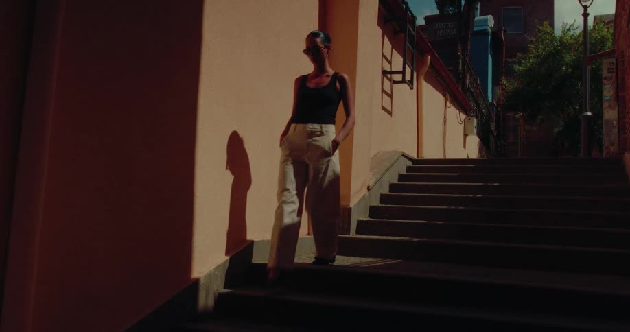A fashionable woman in sunglasses slowly walks down a city staircase, moving from bright sunlight into deep shadow