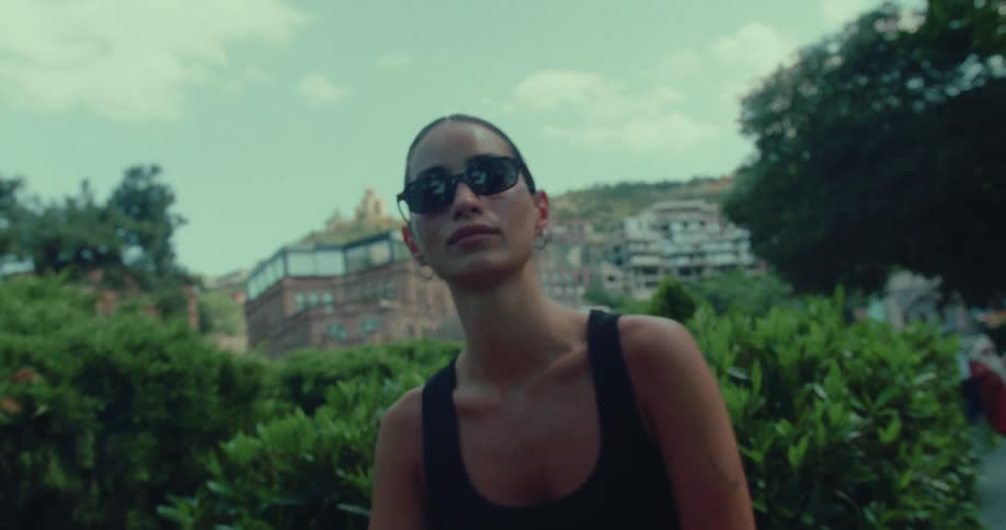 A fashionable woman in sunglasses poses on a park bench as the camera slowly zooms in, with a historic hillside town in the background