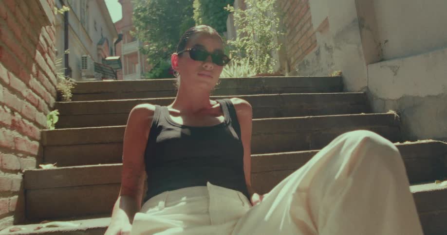 A stylish woman in sunglasses and a black tank top sits on old city steps, enjoying the warm summer sunshine in a slow-motion shot