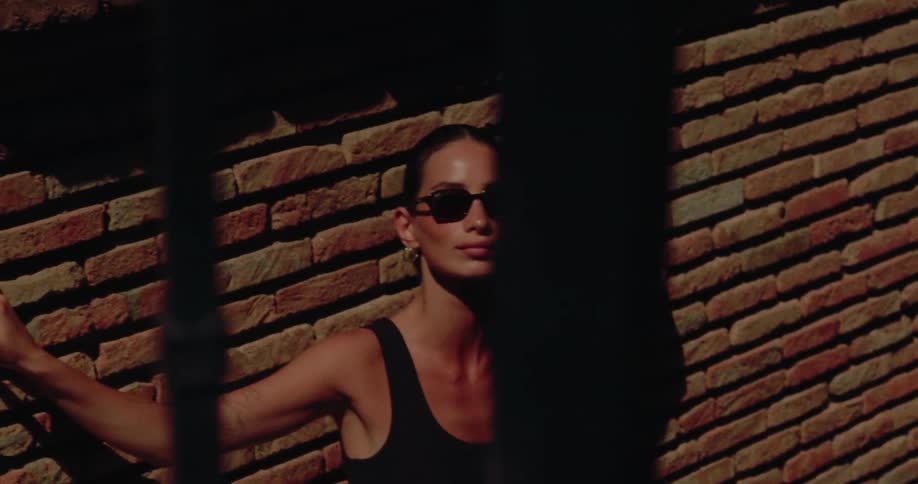 Through the shadows of a fence, a chic woman in sunglasses models against a brick wall in the golden hour light