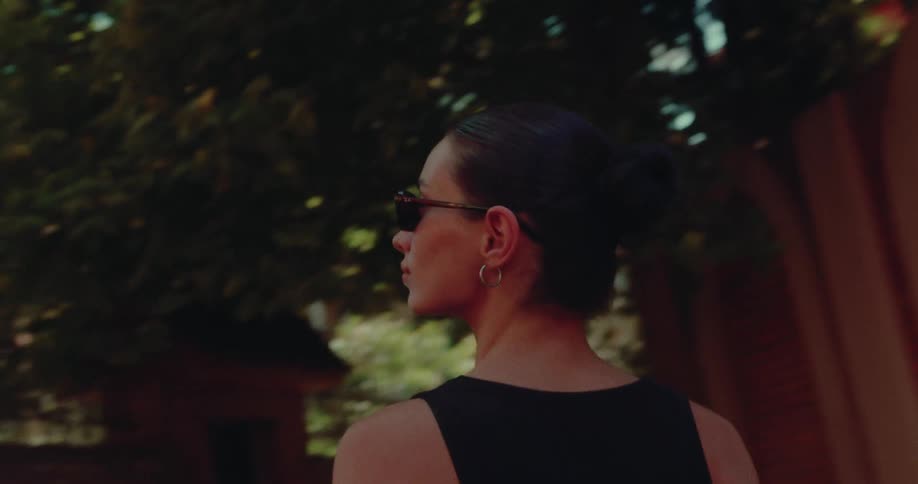 A stylish woman in sunglasses walks down a sunny, tree-lined city street in a cinematic slow-motion follow shot