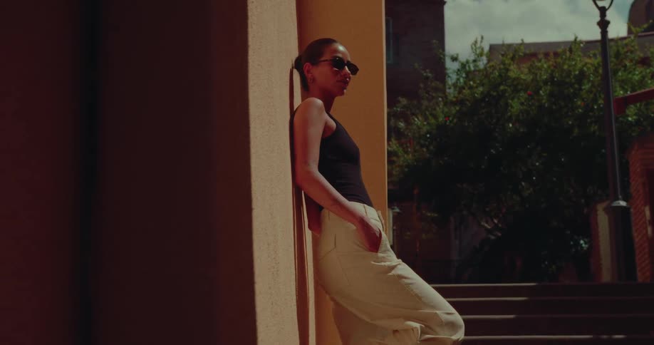 A stylish woman in sunglasses casually leans against a sun-drenched city wall as the camera slowly pans past her