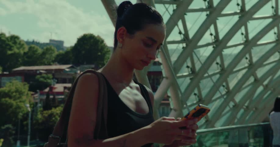 A young woman in a black tank top texts on her smartphone while standing on a modern architectural bridge during the day