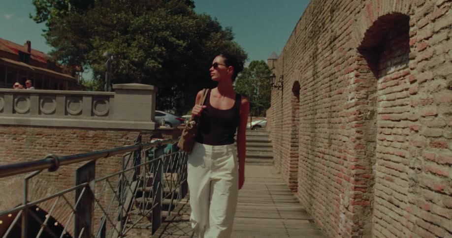 A stylish woman in sunglasses and a tank top enjoys a leisurely walk in slow motion along a historic brick wall on a sunny summer day
