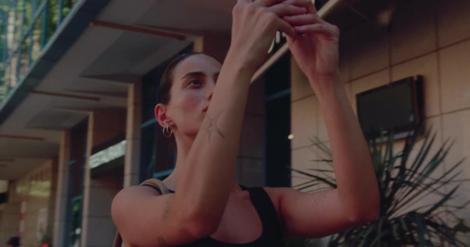 A young woman with tattoos holds her phone up to take a picture while walking down a city street in slow motion