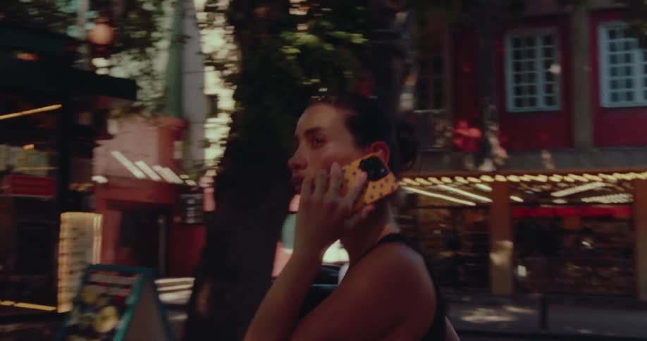 Young woman in a black tank top talks on her smartphone while walking down a sunny city street