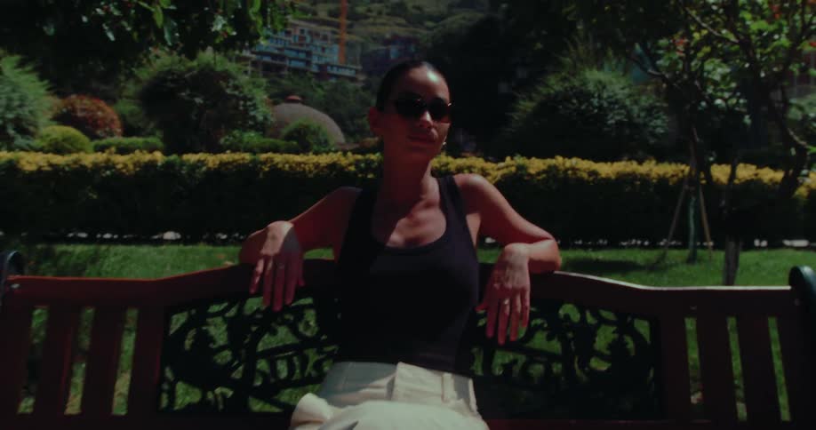 A stylish woman wearing sunglasses sits relaxed on a park bench, enjoying a sunny day in a lush green garden