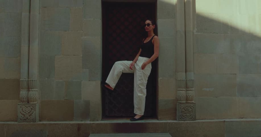 A fashionable woman in sunglasses strikes a stylish pose in a stone doorway with dramatic sunlight and shadows