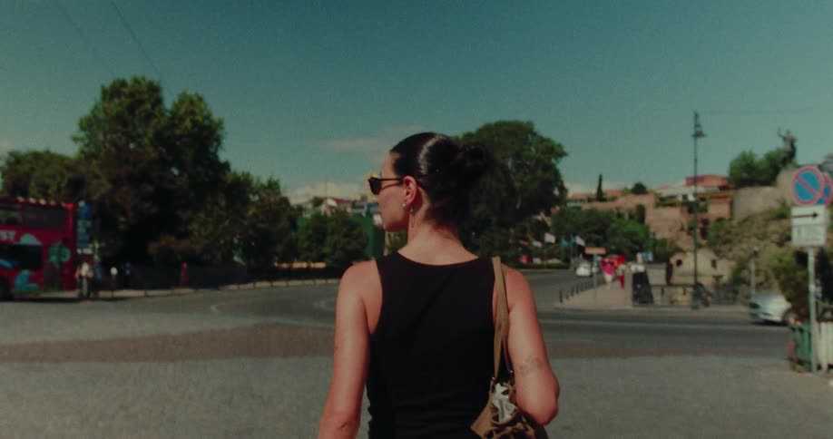 A stylish woman with her back to the camera walks through a sunny, historic European city in slow motion