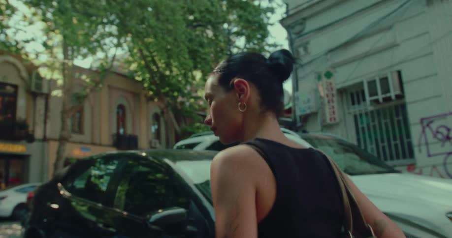 A young woman with dark hair in a bun walks down a sunny, tree-lined city street in slow motion