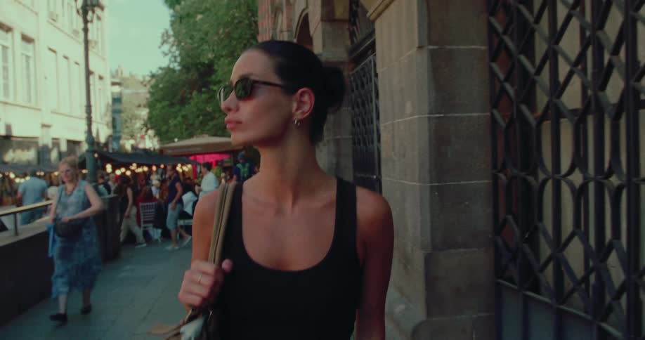 A stylish brunette woman in sunglasses walks down a bustling European city street in slow motion during the golden hour
