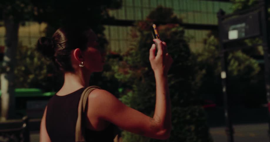 A young woman stands on a sunny city street, holding up her smartphone to capture a photo or video