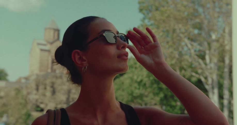 A chic woman takes off her sunglasses while sightseeing on a sunny day with an ancient monastery in the background