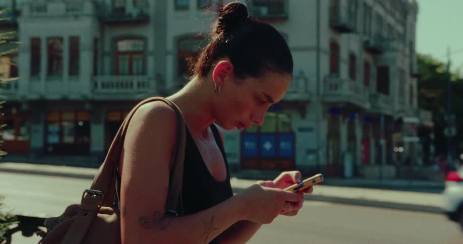 In slow motion, a young woman with her hair in a bun uses her phone to order a ride and then hails it on a sunny city street