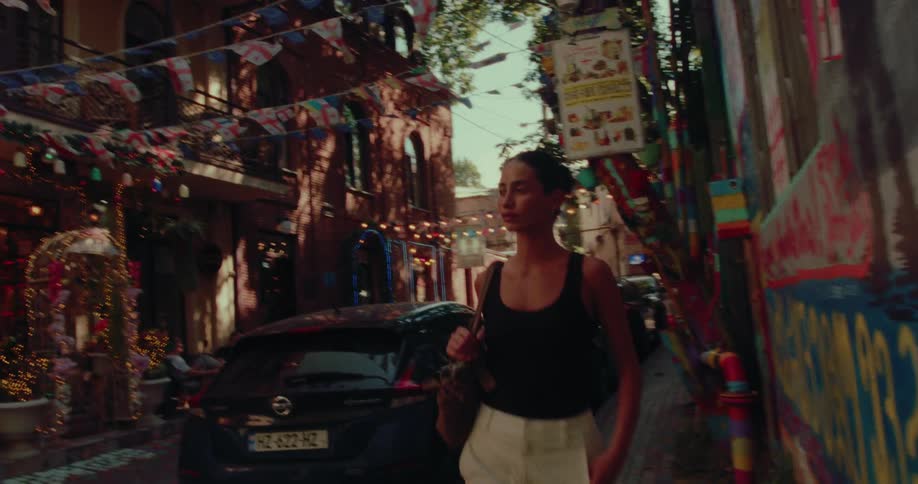 A woman strolls down a festive, narrow cobblestone street adorned with colorful graffiti, string lights, and Georgian flags