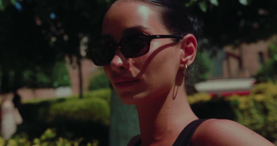 A stylish young woman wearing sunglasses turns to look at the camera in slow motion on a sunny day in a park
