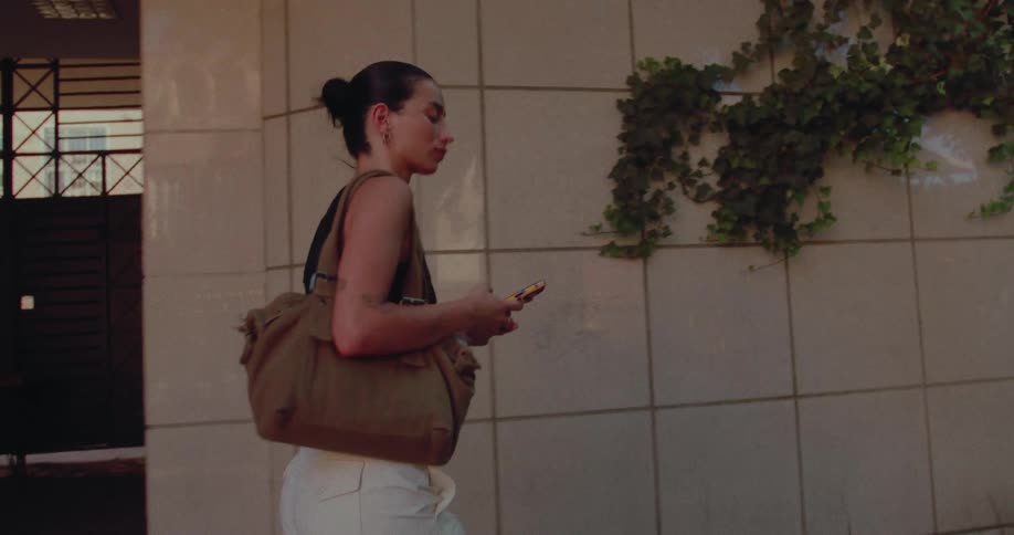 A stylish young woman with her hair in a bun walks down a city street, looking at her smartphone in the warm evening light
