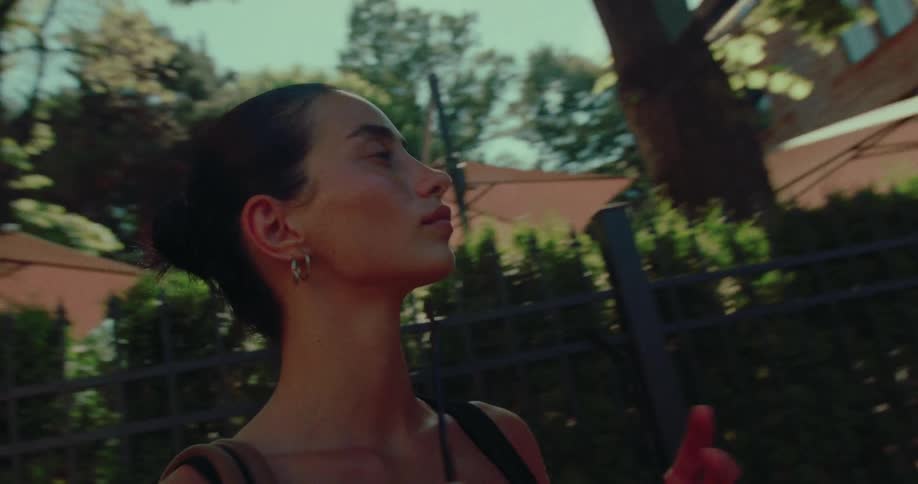 A contemplative woman with dark hair in a bun walks in slow motion along a sunlit, tree-lined city street