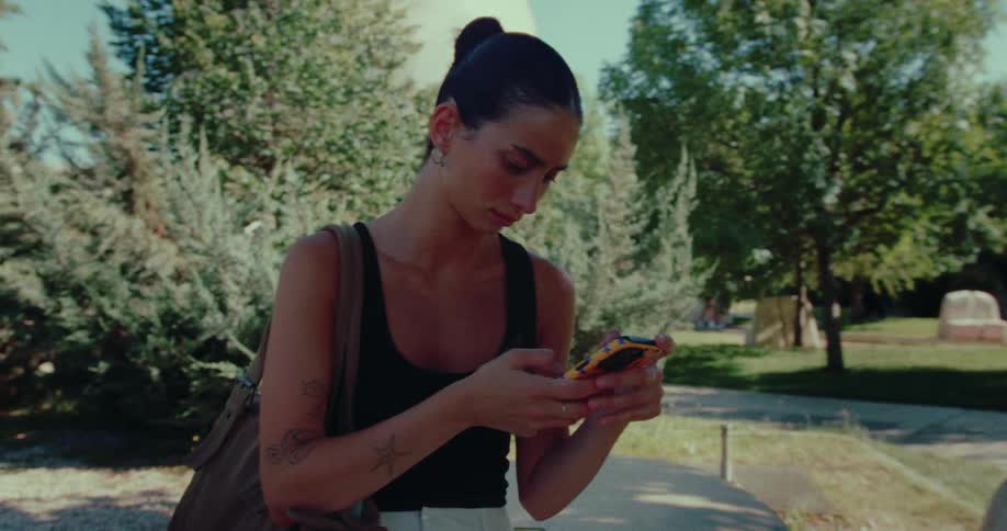 A young woman with tattoos stands in a sunlit park, intently focused on her smartphone as the camera slowly orbits around her