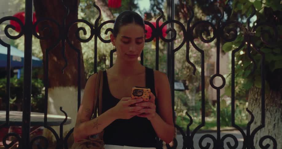A stylish young woman texts on her smartphone while standing in front of an ornate wrought iron gate in a park