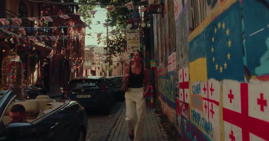 A young woman strolls down a narrow, festive street in Tbilisi, passing a vibrant wall covered in political graffiti and flags