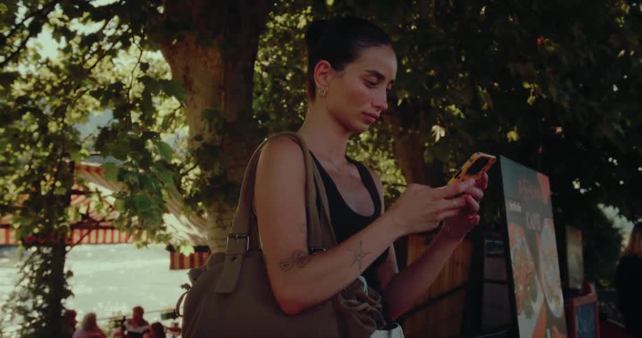 A woman with a tote bag stands under a tree by a sunlit river, engrossed in her smartphone